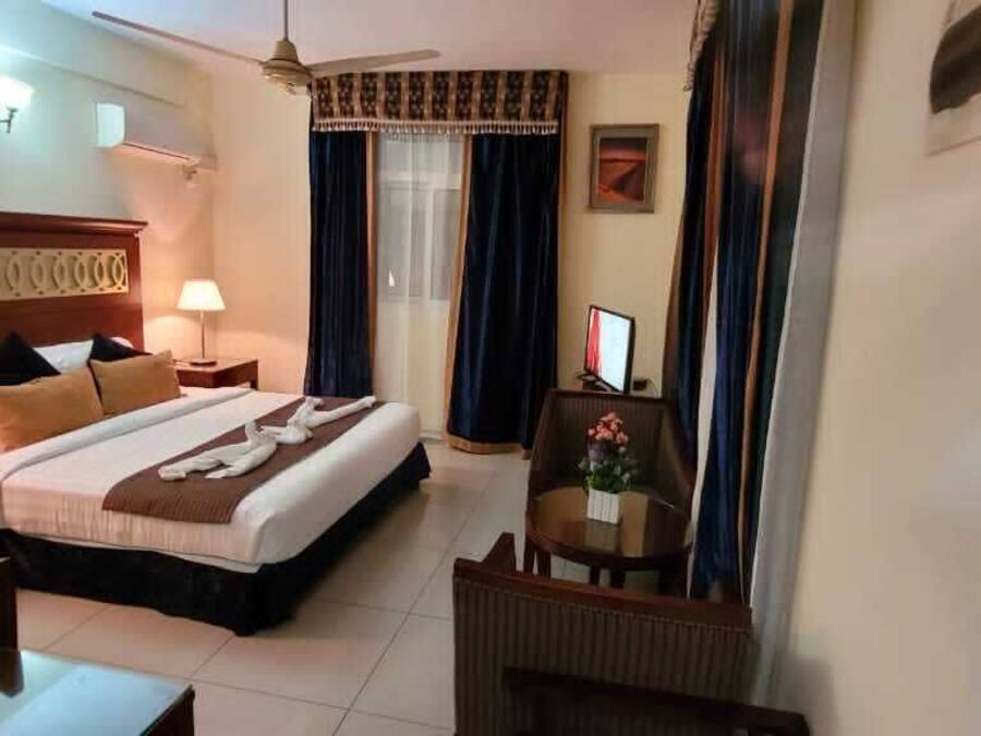 A hotel room with a bed, two bedside tables with lamps, a desk, a chair, a TV, and a ceiling fan.