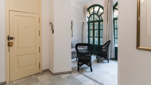 Un couloir avec une porte de couleur crème, un sol à motifs et deux fauteuils en osier près d’une fenêtre aux cadres verts.