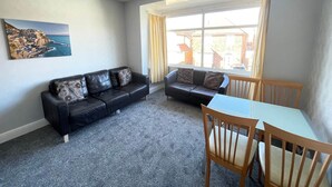 A living room with a black leather sofa, a wooden dining table with chairs, a large window with curtains, and a painting on the wall. - 2 Bedroom Holiday Apartment - Flat 18 (Skegness)