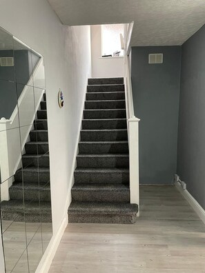 Un escalier avec des marches recouvertes d’une moquette sombre et des rampes blanches, menant à un palier doté d’une petite fenêtre.