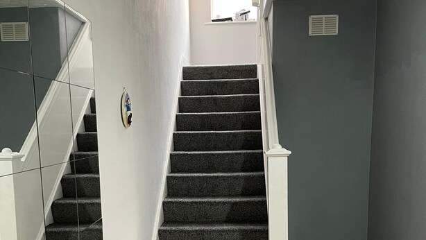 Un escalier avec des marches recouvertes d’une moquette sombre et des rampes blanches, menant à un palier doté d’une petite fenêtre.