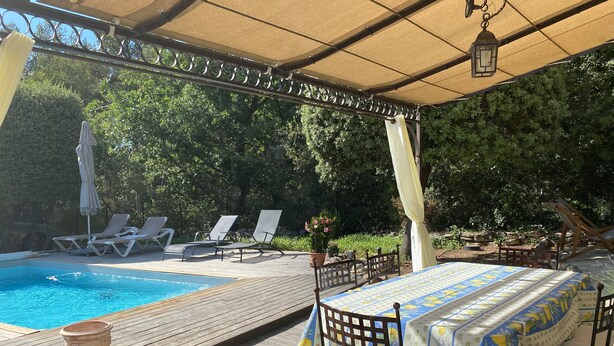 A poolside area with a wooden deck, lounge chairs, a table with a tablecloth, and a pergola with a canopy.