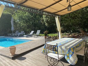 Un espace aménagé au bord de la piscine, avec une terrasse en bois, des chaises longues, une table recouverte d’une nappe et une pergola dotée d’un auvent.