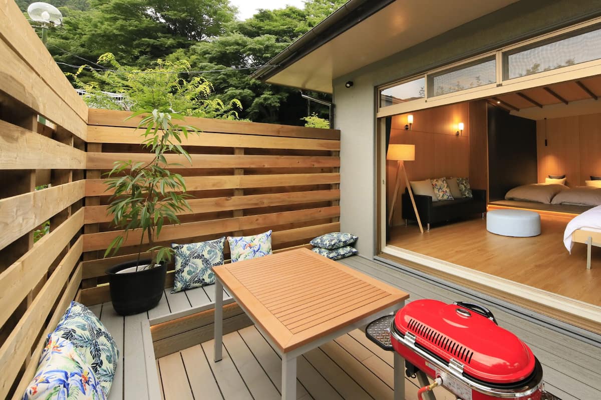 Een houten terras met een tafel, stoelen, een rode barbecue en uitzicht op een slaapkamer met een bed en nachtkastje.