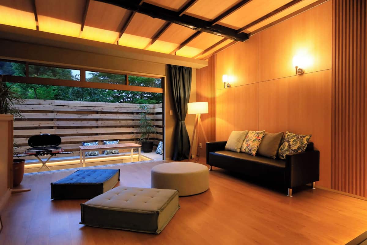 Een moderne woonkamer met een houten vloer, een zwarte bank met bloemenkussens, een rond poefje, een kleine tafel met een lamp en uitzicht op groen door grote ramen.