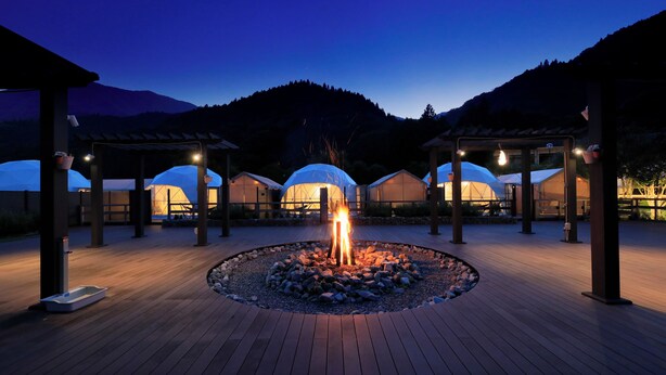 Une terrasse en bois dotée d’un foyer central, entourée de structures abritées et avec une chaîne de montagnes en arrière-plan.