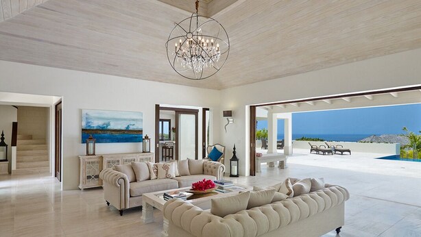 A spacious living room with a large sofa, a coffee table, and a chandelier. There is a painting on the wall and a view of the ocean through large windows. - Infinity House | Ultra-Luxury Sea View Estate in Calijanda, Barbados (Holetown)