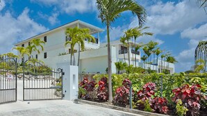 A two-story building with a gated entrance, surrounded by palm trees and red-leafed plants. - Infinity House | Ultra-Luxury Sea View Estate in Calijanda, Barbados (Holetown)
