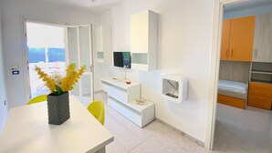 A modern hotel room with a white color scheme, a ceiling fan, a flat-screen TV, a small seating area with a table and chairs, and a view through an open door.
