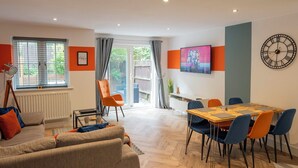 A modern living room with a sofa, a dining table with chairs, a TV mounted on the wall, and a large clock. - Stylish, City Centre 3-bed House With Parking (Canterbury)