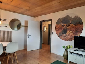 A modern room with a wooden ceiling, a round mirror, a wall-mounted TV, a dining area with chairs, and a door leading to another room.