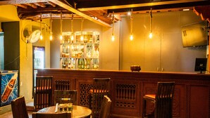 Een bar met houten stoelen en tafels, een barvloer met flessen en hangende lampen.