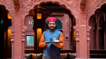 A person in traditional attire holding a tray with a glass and a folded cloth, standing in front of an intricately carved architectural structure.