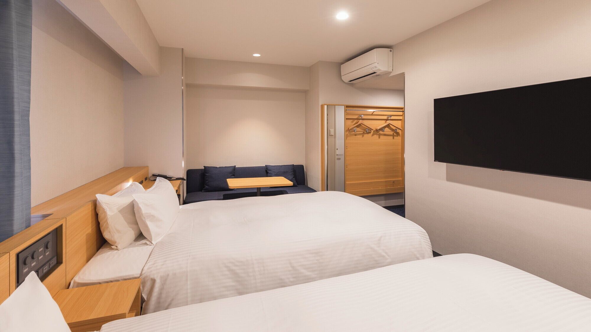 Ein Hotelzimmer mit zwei Betten, einem Holz-Kopfteil, einem Nachttisch, einem an der Wand befestigten Fernseher und einer Klimaanlage.