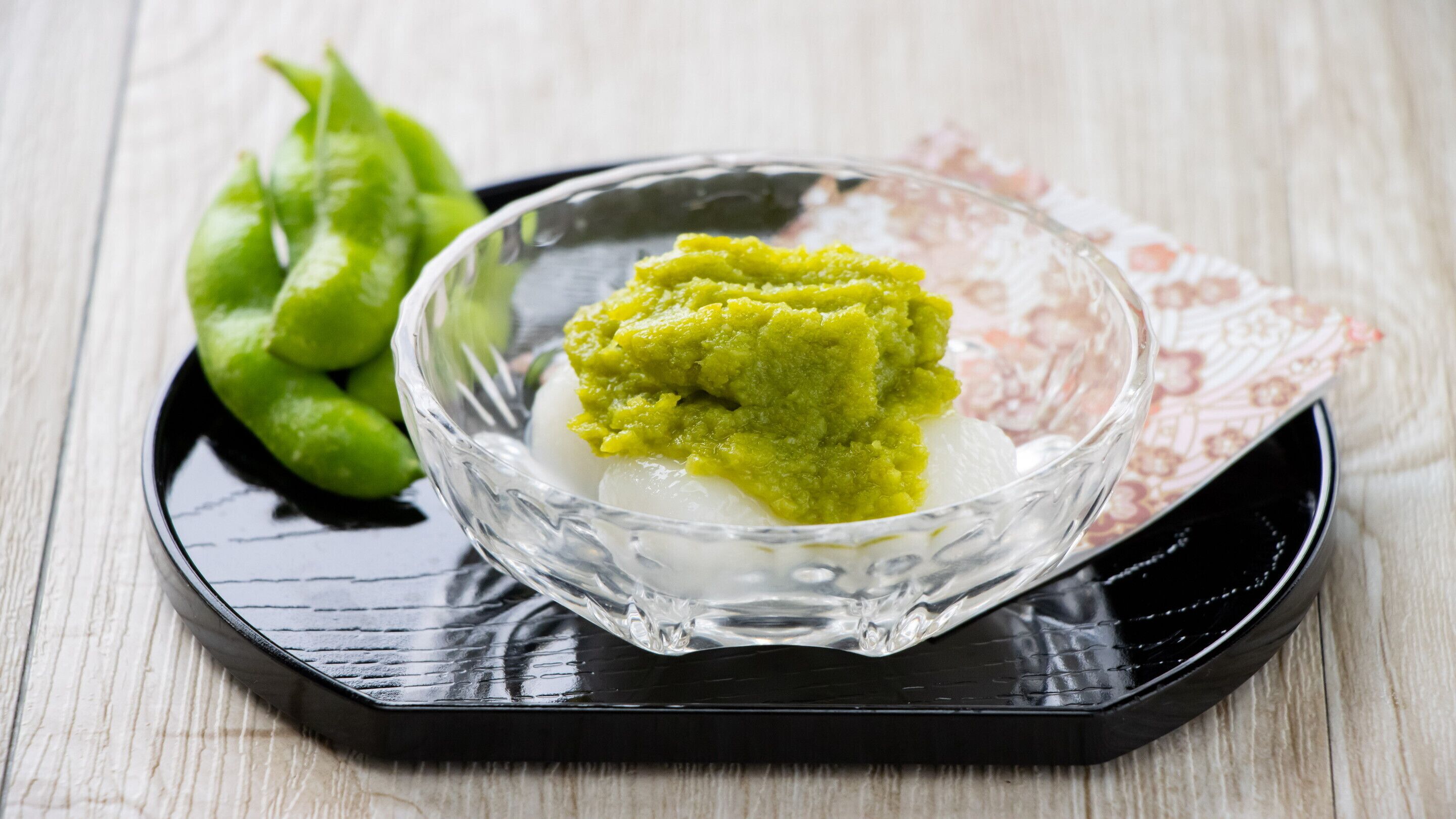 A clear glass bowl containing a white spherical food item with a green topping, placed on a black tray with a floral-patterned napkin underneath.