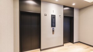 Un ascenseur moderne doté d’un panneau d’affichage numérique, encadré par des portes d’ascenseur de couleur sombre, dans un couloir aux murs de couleur claire et au revêtement de sol en bois.