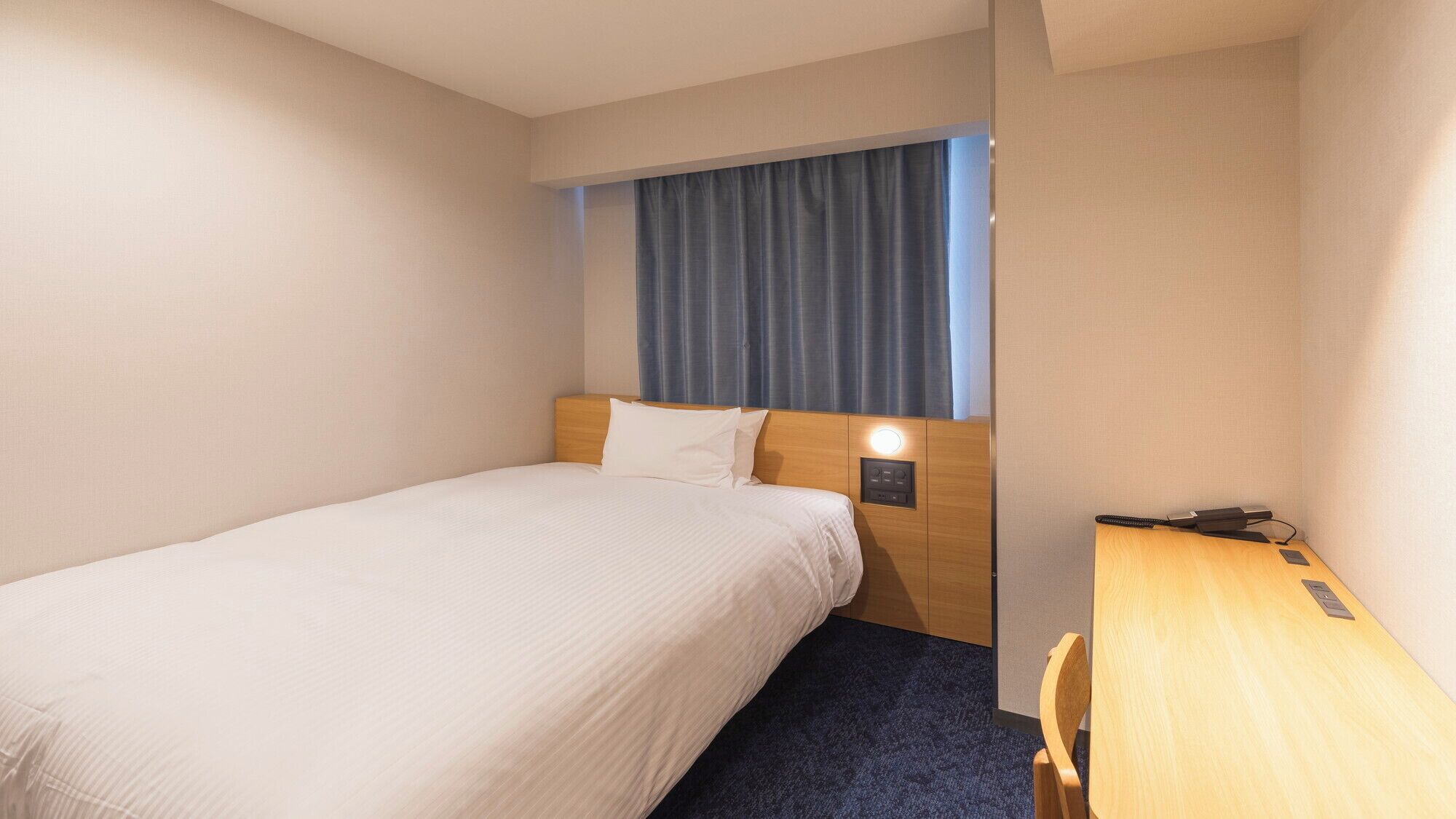 Ein Hotelzimmer mit einem Einzelbett, einem Schreibtisch, einem Nachttisch, einem Fenster mit Vorhängen und einer Wandleuchte.