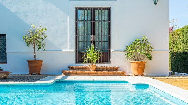 A poolside view of a building with a large window, potted plants, and a clear blue pool. - Holiday Home "Casa Rural La Teja" with Pool, Garden and Wi-Fi (Marchena)