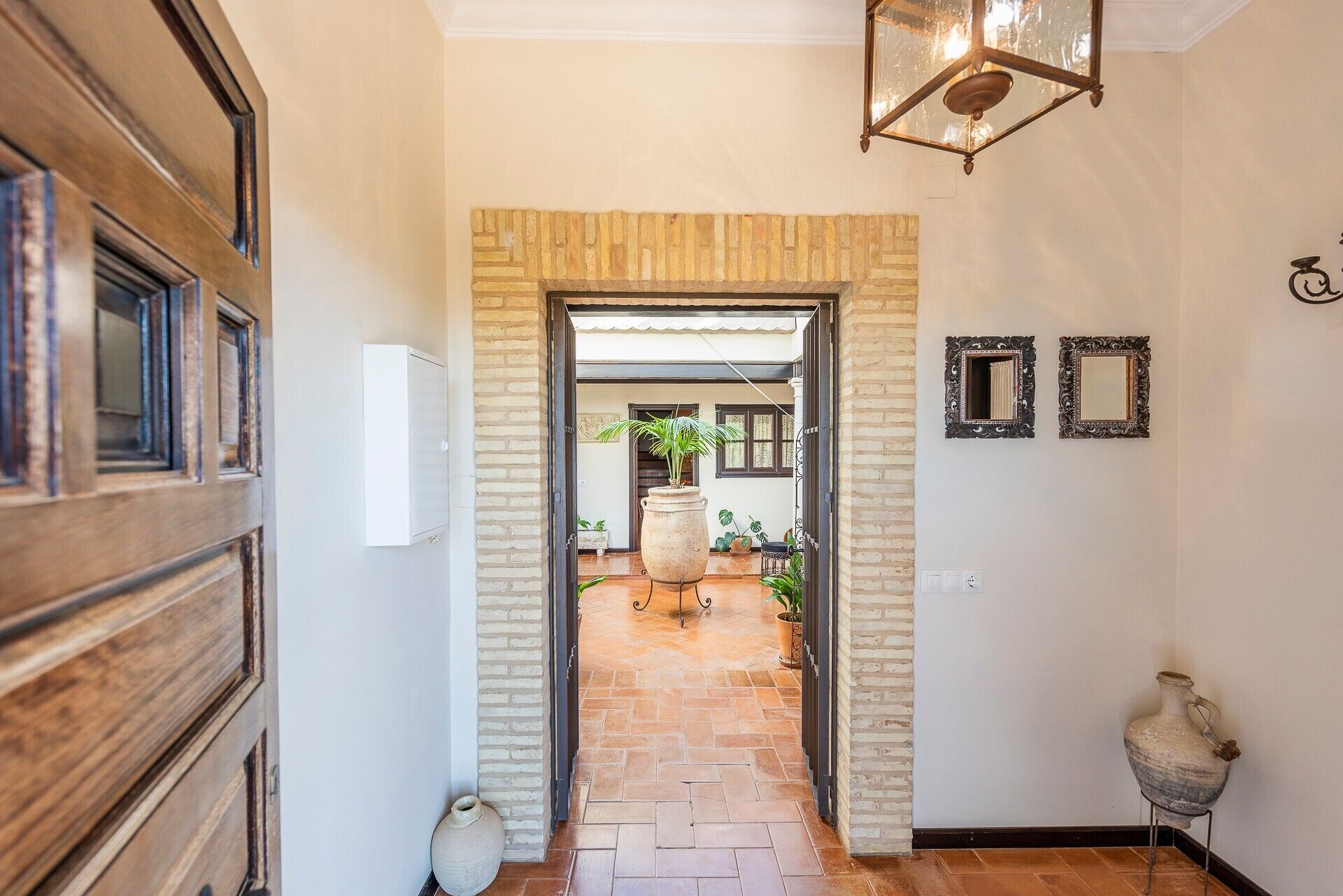 Un pasillo con una puerta de madera, un muro de ladrillo, una maceta grande y dos espejos decorativos.