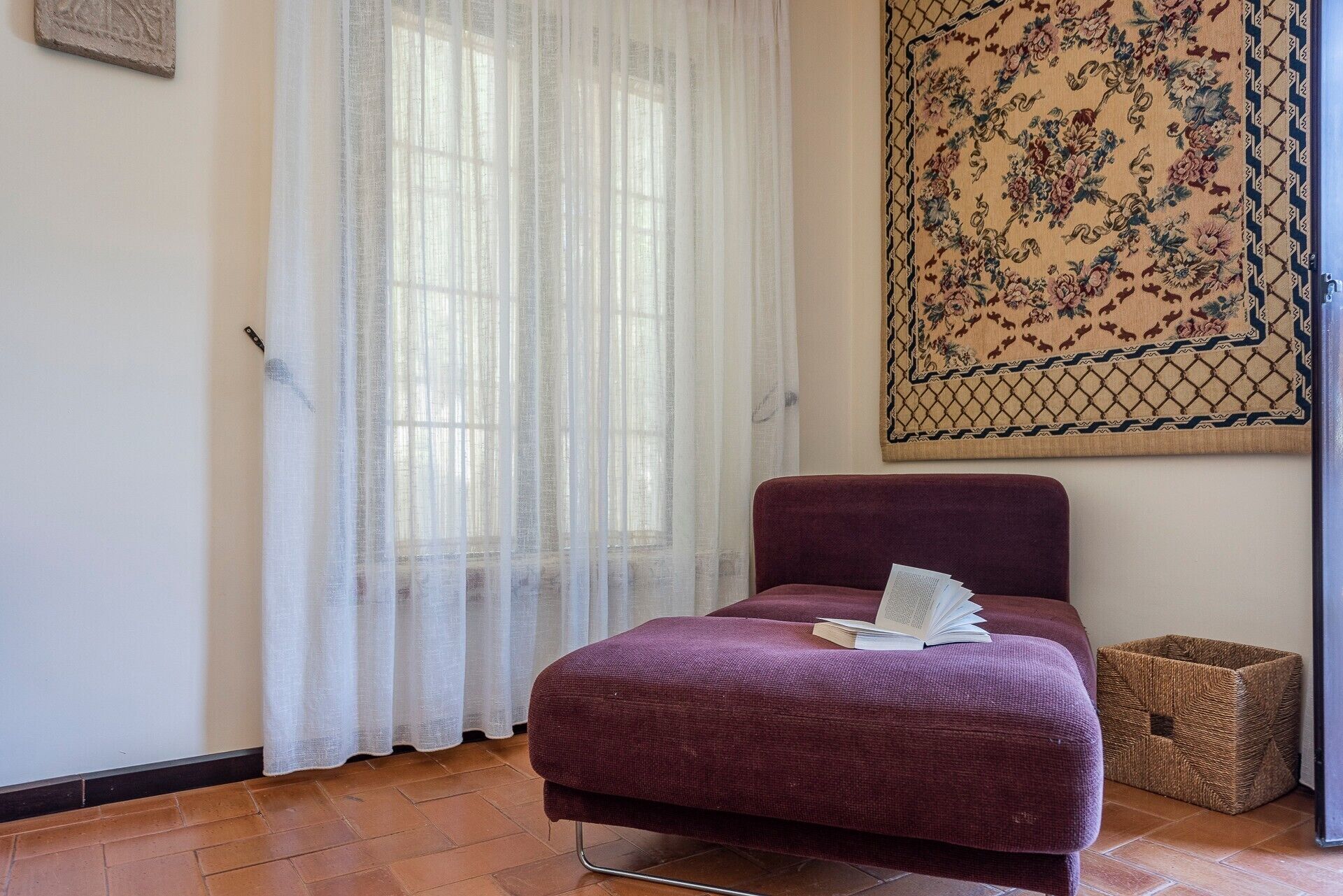 Ein Zimmer mit einem Bett, einem Fenster mit durchscheinenden Vorhängen, einem gewebten Korb und einem Wandteppich.