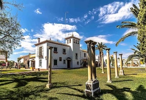 Un edificio blanco con una torre central, rodeado de un césped cuidado y palmeras.