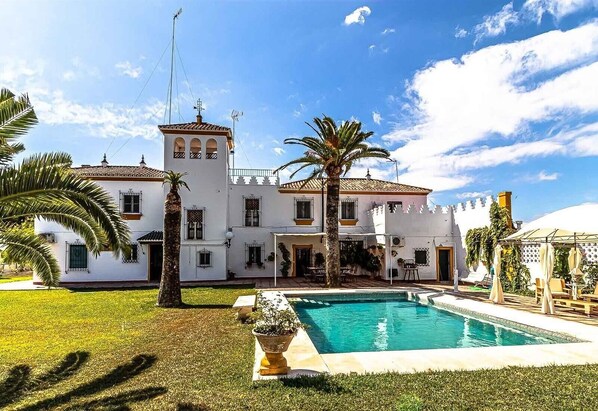 Un edificio blanco grande con piscina, palmeras y un cielo azul despejado.