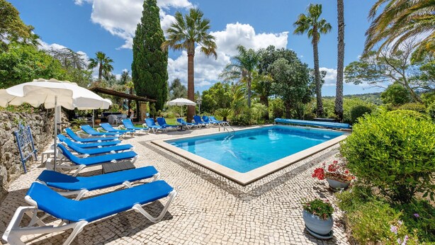 A swimming pool with blue lounge chairs and umbrellas, surrounded by greenery and palm trees. - Quinta das Achadas - Jasmine in Lagos with Shared Pool, Nature & Breakfast (Lagos)