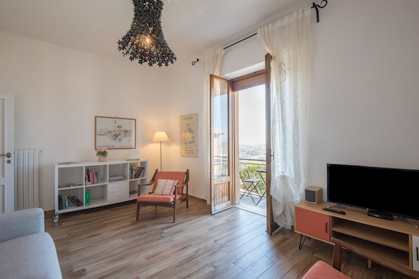Ein Wohnzimmer mit einer Couch, einem Sessel, einem Fernseher, einem Bücherregal und einem Balkon mit Blick.