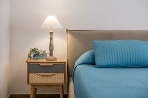 Ein ordentlich bezogenes Bett mit blauer Decke, eine Nachttischlampe und ein kleiner Nachttisch aus Holz mit Schublade und Dekoration.