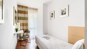 Un dormitorio pequeño y ordenado con una cama individual, una silla, una mesita con una planta y dos cuadros en la pared.