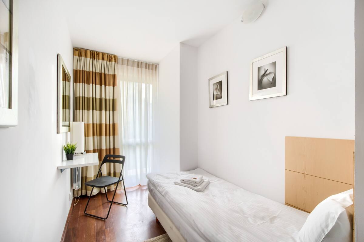 Un dormitorio pequeño y ordenado con una cama individual, una silla, una mesita con una planta y dos cuadros en la pared.