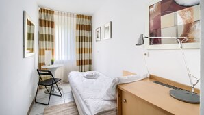Un dormitorio pequeño y ordenado con una cama individual, un escritorio con silla, una mesita de noche, una lámpara y una ventana con cortinas.