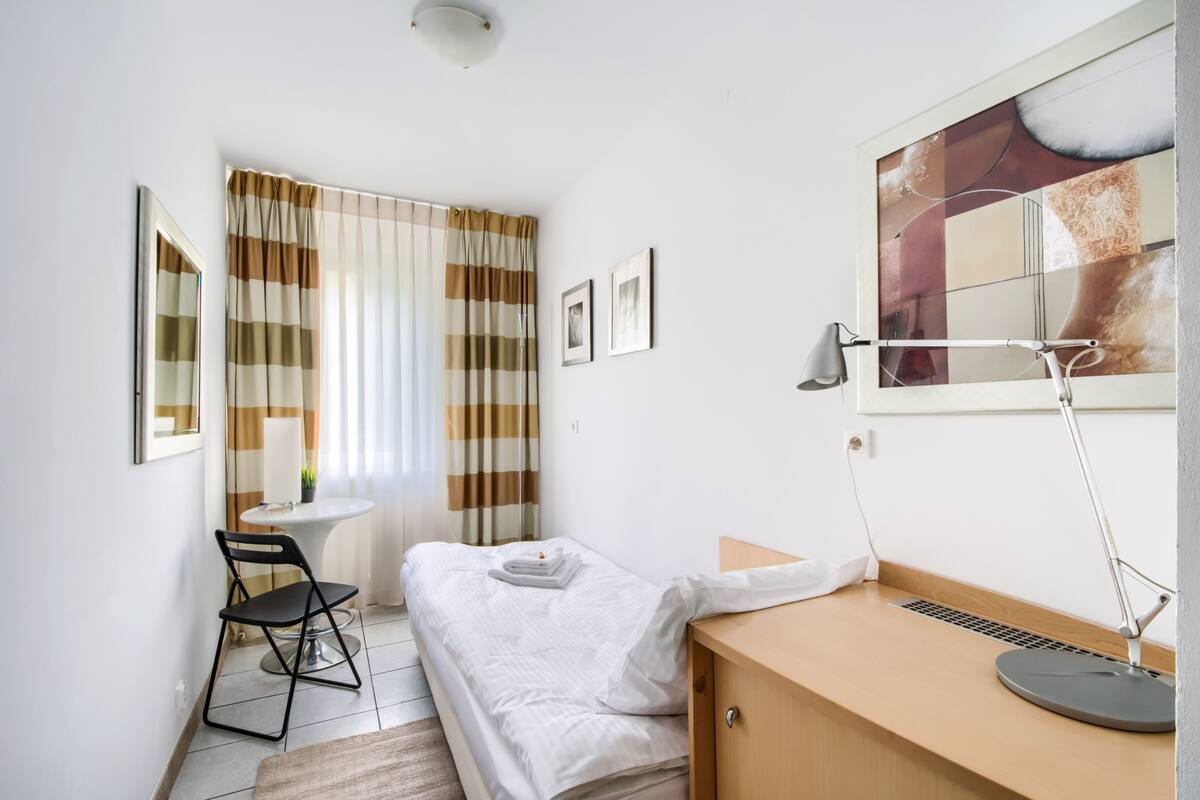 Un dormitorio pequeño y ordenado con una cama individual, un escritorio con silla, una mesita de noche, una lámpara y una ventana con cortinas.