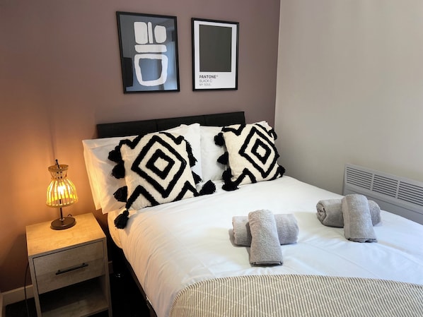 A neatly made bed with two black and white patterned pillows, a bedside lamp, and three folded towels on the bed. - The Gateway Apartment 4 (Blackpool)