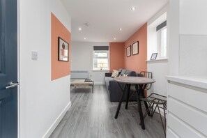A modern living space with a sofa, dining table, and chairs. There is a window with a view, a door, and a light switch on the wall.