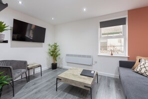 A modern living room with a flat-screen TV, a coffee table, a sofa, and a window with a view. - The Gateway Apartment 4 (Blackpool)