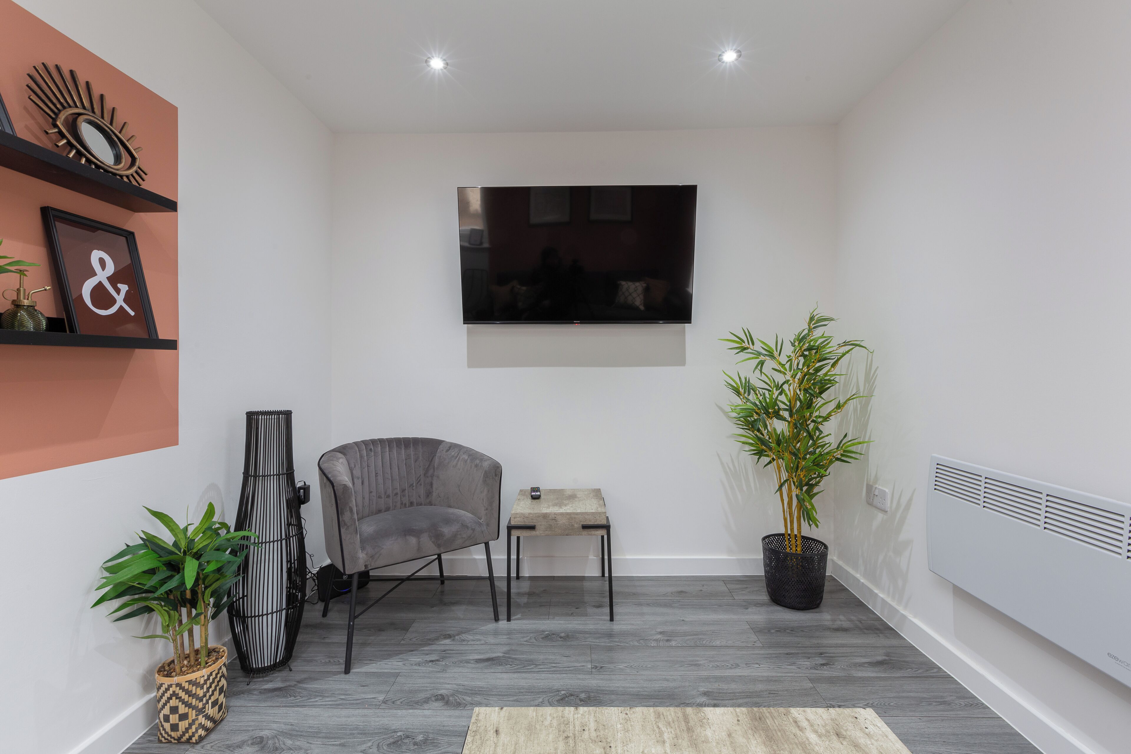 Un salon moderne avec un fauteuil gris, une petite table, une grande plante en pot, un téléviseur fixé au mur et un miroir décoratif.