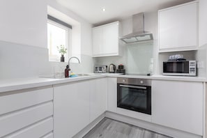 A modern kitchen with white cabinetry, a built-in oven, a microwave, and a window with a plant. - The Gateway Apartment 4 (Blackpool)