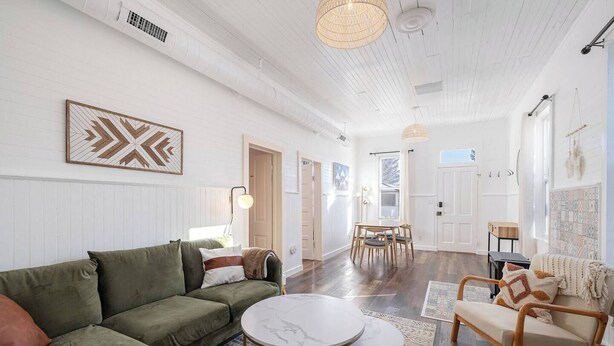 A spacious living room with a green sofa, a round coffee table, and a wooden chair. There is a dining area with a table and chairs, and a large window providing natural light.