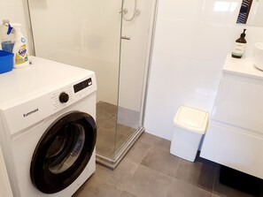 A modern laundry room with a front-loading washing machine, a glass shower enclosure, and a small white sink. - Earthy Mix M6 Studio Apartment near Kings Park (West Perth)