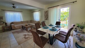 A living room with a sofa, coffee table, and dining table set with chairs. There is a ceiling fan, a window with curtains, and a view of greenery outside. - Villa - Sleeps 6, Free Parking & Heated Pool (Paralimni)