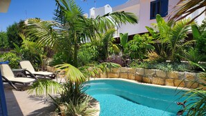 Une piscine entourée de chaises longues, de plantes tropicales et d’un mur en pierre.