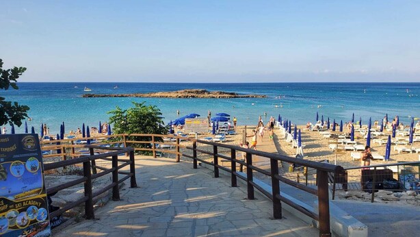 Une plage avec une promenade en bois menant à l’eau, des parasols bleus et un panneau indicateur.