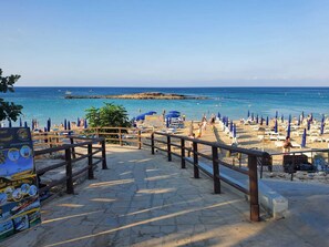 A beach with a wooden walkway leading to the water, blue umbrellas, and a signboard. - Charming 3-bed Villa in Protaras With Heated Pool (Paralimni)