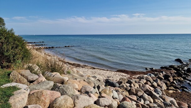 A rocky beach with clear blue water and a distant sailboat. - Welcome to apartment 58 in the house "Berlin". (Großenbrode)
