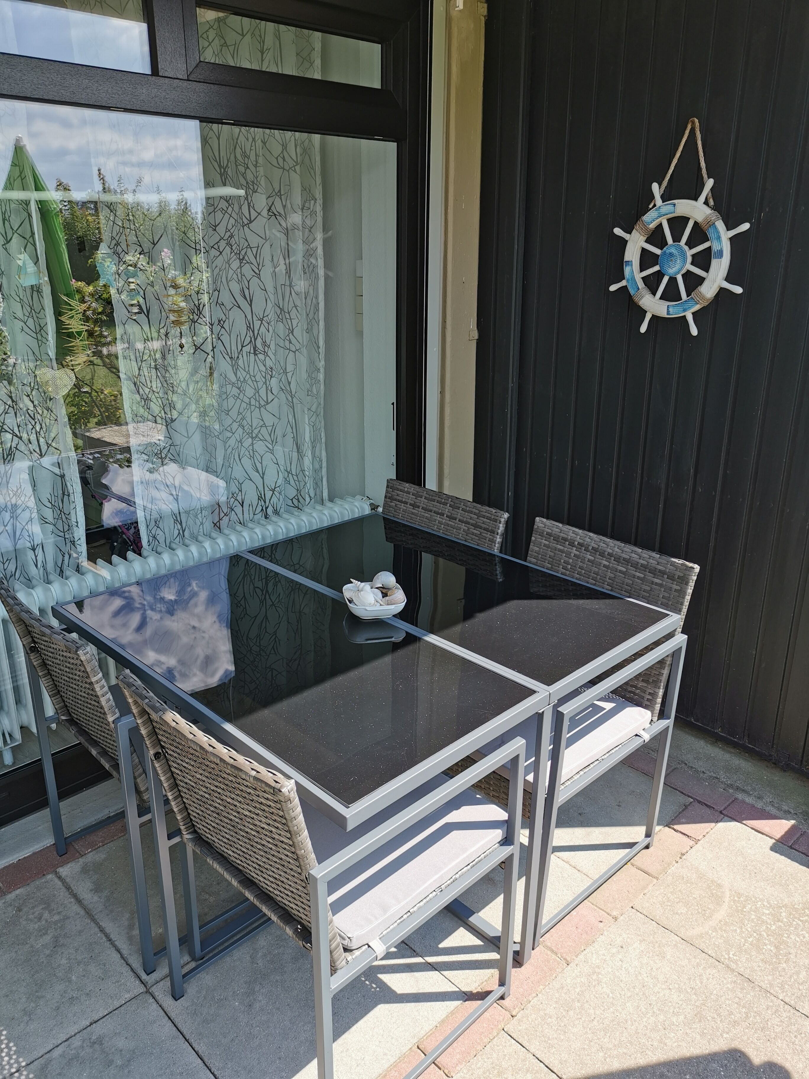 An outdoor patio with a glass-top table and wicker chairs, a decorative wheel, and a view of greenery through a patterned window.