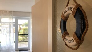 A doorway with a 'WELCOME ON BOARD' sign, a glass door leading outside, and a view of greenery through the window. - Welcome to apartment 58 in the house "Berlin". (Großenbrode)