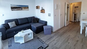 A modern living room with a dark-colored sofa, a white coffee table, and a gray ottoman. There is a wooden door leading to a hallway with a white cabinet and a small table. - Welcome to apartment 58 in the house "Berlin". (Großenbrode)