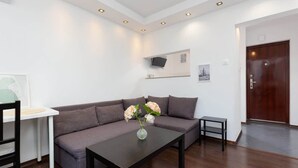 Una sala moderna con un sofá marrón oscuro, una mesita de centro con un jarrón de flores, una mesa blanca con una silla y una puerta de madera.