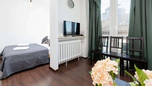 Habitación de hotel con cama, televisión, radiador, ventana con cortinas y mesa con sillas.